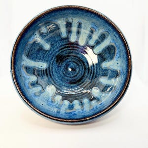 May include: A handmade ceramic bowl with a deep blue glaze and a textured interior. The bowl features a dark brown rim and a unique pattern of raised, light blue shapes. The bowl's interior has a spiral design.