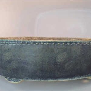 May include: A round, low-profile ceramic bonsai pot with a dark, mottled blue-grey finish. The upper rim features a raised, beaded texture, and the base is supported by three small, decorative feet with a light-colored, contrasting edge.