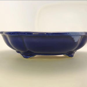 May include: A deep blue ceramic bowl with a scalloped rim and a glossy finish. The bowl features a rounded, lobed body supported by three small, short feet. A small area of lighter blue or white glaze is visible on the upper right edge.