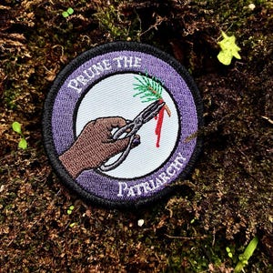 May include: A circular embroidered patch with a purple border and white center. It features a brown hand holding silver shears cutting a green pine branch, with red liquid dripping from the cut. The text reads PRUNE THE PATRIARCHY.