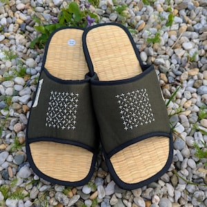 Può includere: Un paio di pantofole verdi e beige con un plantare in paglia intrecciata e una tomaia in tessuto verde scuro. La tomaia presenta un disegno ricamato bianco e un bordo nero. Le pantofole sono appoggiate su un letto di piccole pietre.