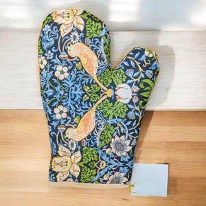 Puede incluir: Un guante de cocina colorido con un estampado floral y de pájaros. El guante presenta un fondo azul oscuro con diseños florales y de pájaros en azul claro, amarillo y verde. Los bordes están ribeteados con tela amarilla. Se adjunta una etiqueta blanca en blanco.