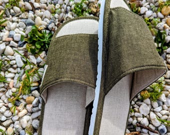 Könnte beinhalten: Ein Paar offene Hausschuhe mit olivgrünem Stoffobermaterial und hellbeigen Stoffinnensohlen. Die Hausschuhe verfügen über eine weiße Gummisohle mit strukturiertem Rand, die einen Kontrast zu den gedeckten grünen und neutralen beigen Farbtönen bildet.