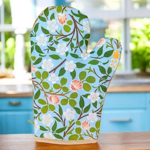 Può includere: Un guanto da forno azzurro con un motivo floreale di fiori bianchi, boccioli arancioni e foglie verdi su rami marroni. Il guanto è in tessuto e ha un bordo beige. Un anello è attaccato al lato.