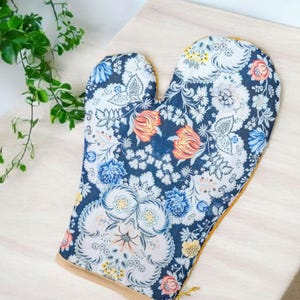 Peut inclure: Un gant de cuisine floral avec un fond bleu marine et un motif coloré de fleurs blanches, orange et bleues. Le gant a une bordure beige et est conçu pour la cuisine.