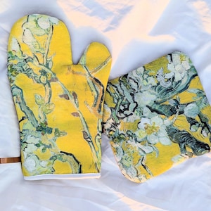 Puede incluir: Un guante de cocina amarillo y un salvamanteles cuadrado a juego con un diseño floral en tonos verdes, blancos y marrones. El guante tiene un lazo marrón para colgar y ribete blanco. El juego es para la cocina.