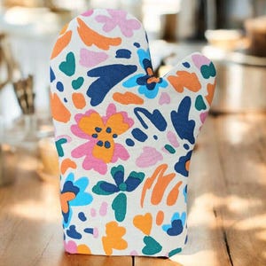 Op de afbeelding: Een kleurrijke ovenwant met een bloemenpatroon. De want heeft een witte achtergrond met oranje, roze, blauwe en groene bloem- en bladvormen. Ontworpen voor gebruik in de keuken.