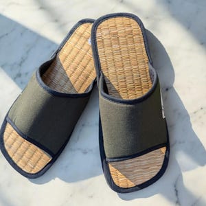Può includere: Un paio di pantofole intrecciate verdi e beige. Le pantofole hanno una tomaia in tessuto verde scuro e un plantare intrecciato beige. I bordi sono rifiniti con un tessuto blu scuro. Le pantofole sono progettate per uso interno.