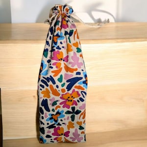 Peut inclure: Un sac en tissu beige avec un motif floral aux couleurs vives : orange, rose, bleu et vert. Le sac est noué en haut avec un cordon blanc. Le sac est posé sur une surface en bois.