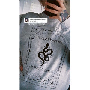 Könnte beinhalten: Graues Sweatshirt mit einem schwarzen Schlangen- und Blumendesign in einem rechteckigen Rahmen. Der Rahmen enthält dekorative Elemente und Text. Die Wörter "PATHOLOGICAL" und "PEOPLE PLEASER" sind sichtbar. Das Sweatshirt hat auch den Text "@pumpkinavis.design".