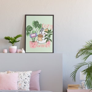 May include: A framed print featuring a cartoon cat holding a coffee cup and a game controller, surrounded by plants. The text "Cozy Gamer" is displayed in pink. The print is set above a bed with pink pillows and a gray comforter.