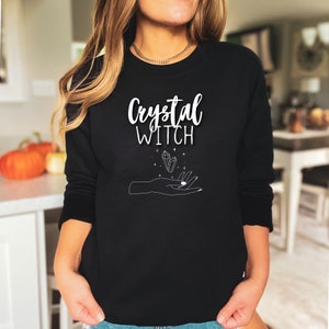 May include: Black sweatshirt with white text that reads "Crystal Witch" and a graphic of a hand holding a crystal.