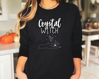 Könnte beinhalten: Schwarzer Pullover mit weißem Text "Crystal Witch" und einer Grafik einer Hand, die einen Kristall hält.