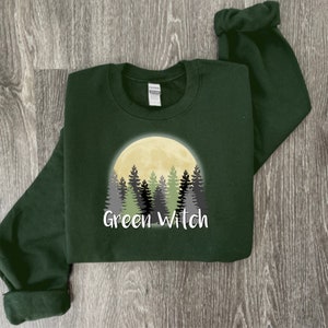 May include: A dark green sweatshirt with a graphic of a yellow moon and a forest of green and gray trees. The text "Green Witch" is printed below the graphic.