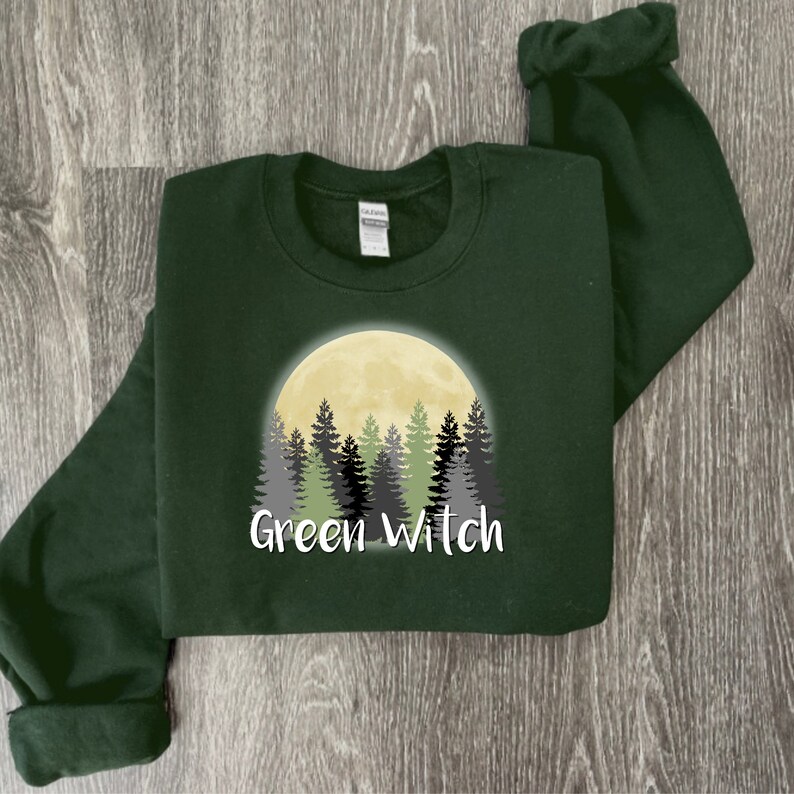May include: A dark green sweatshirt with a graphic of a yellow moon and a forest of green and gray trees. The text "Green Witch" is printed below the graphic.