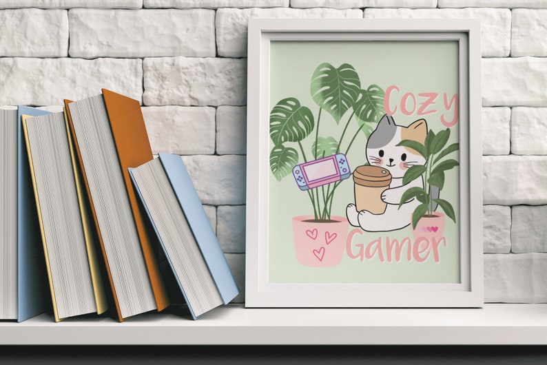 May include: A framed print with a cat illustration holding a coffee cup, with the text "Cozy Gamer." The image includes a video game console and potted plants. Books are visible on a shelf to the left.