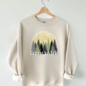 May include: A beige sweatshirt with a graphic design of a full moon and a forest silhouette. The text "Green Witch" is printed below the image.