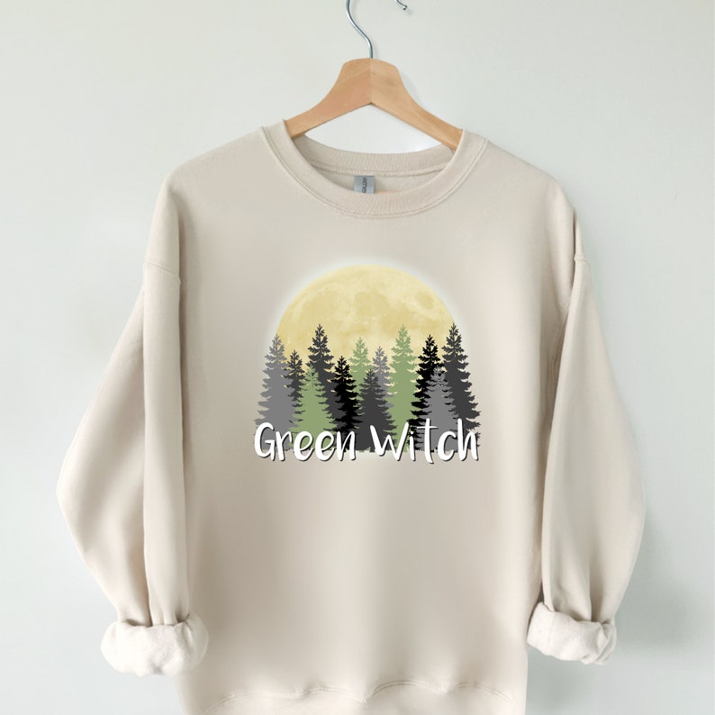 May include: A beige sweatshirt with a graphic design of a full moon and a forest silhouette. The text "Green Witch" is printed below the image.