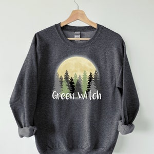 May include: A dark gray sweatshirt with a graphic of a yellow moon behind a forest of green trees. The text "Green Witch" is printed below the moon.