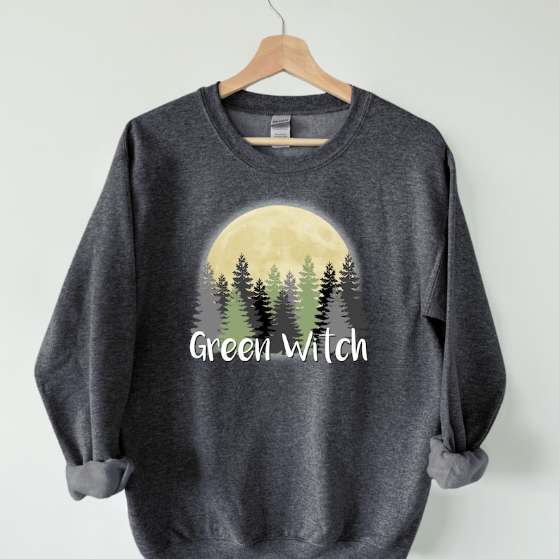 May include: A dark gray sweatshirt with a graphic of a yellow moon behind a forest of green trees. The text "Green Witch" is printed below the moon.