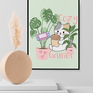 May include: A framed print featuring a cartoon cat holding a coffee cup and a game console, surrounded by potted plants. The words "Cozy Gamer" are written in pink. The background is a light green color.