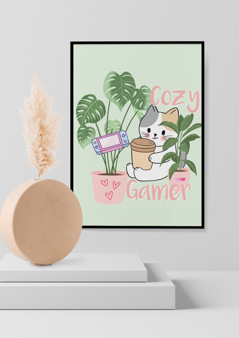 May include: A framed print featuring a cartoon cat holding a coffee cup and a game console, surrounded by potted plants. The words "Cozy Gamer" are written in pink. The background is a light green color.