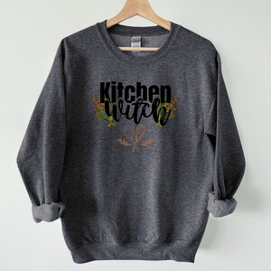 K&ouml;nnte beinhalten: Ein dunkelgrauer Pullover mit dem Text "Kitchen Witch" in schwarzen Buchstaben mit einem Blumendesign und einem Paar gekreuzter K&uuml;chenutensilien.
