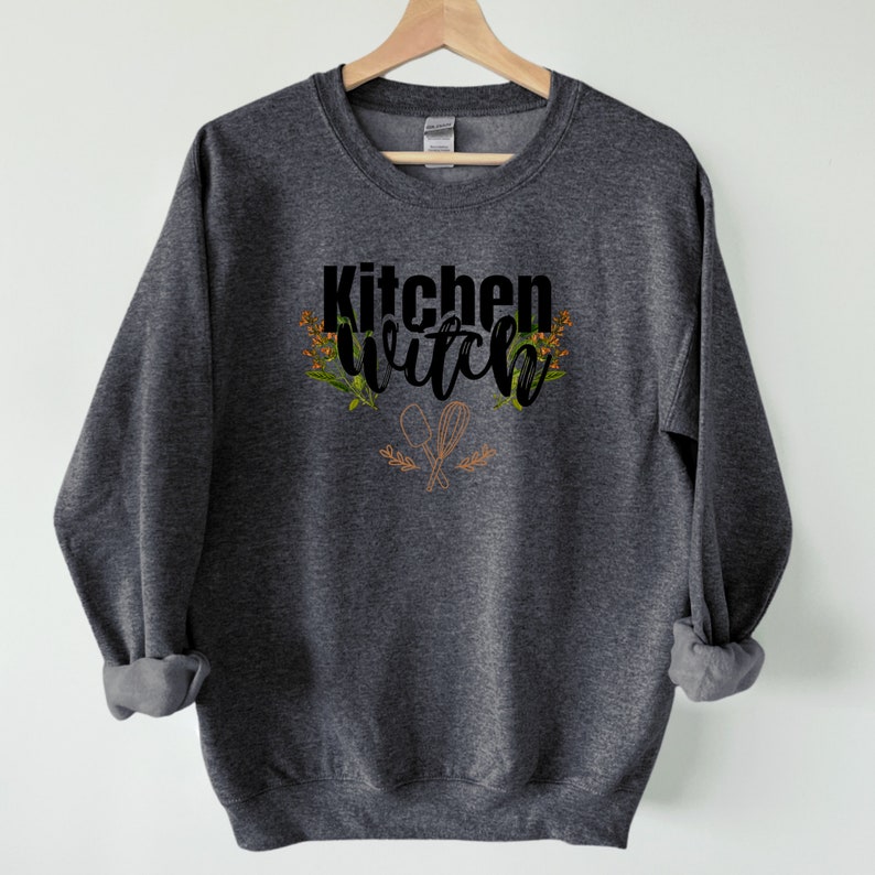 K&ouml;nnte beinhalten: Ein dunkelgrauer Pullover mit dem Text "Kitchen Witch" in schwarzen Buchstaben mit einem Blumendesign und einem Paar gekreuzter K&uuml;chenutensilien.