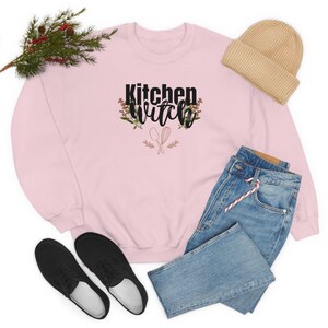 K&ouml;nnte beinhalten: Ein rosa Sweatshirt mit dem Text "Kitchen Witch" in schwarzen Buchstaben mit einem floralen Design und gekreuzten K&uuml;chenutensilien. Das Sweatshirt liegt auf einem wei&szlig;en Hintergrund mit einem Paar schwarzer Turnschuhe, einer blauen Jeans und einer beigefarbenen M&uuml;tze.
