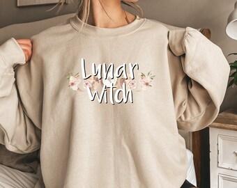 Könnte beinhalten: Ein beiger Pullover mit dem Aufdruck "Lunar Witch" auf der Vorderseite. Der Text ist von einem floralen Design in Rosa- und Brauntönen umgeben.