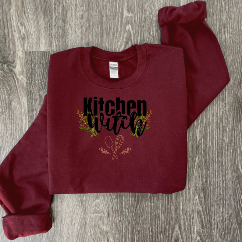 K&ouml;nnte beinhalten: Ein weinroter Pullover mit dem Text "Kitchen Witch" in schwarzen Buchstaben. Der Text ist von kleinen Blumenmustern und einem Paar gekreuzter K&uuml;chenutensilien umgeben.