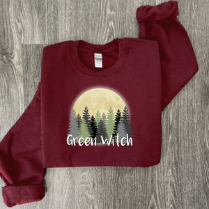 May include: A maroon sweatshirt with a graphic of a full moon and a forest silhouette. The text "Green Witch" is printed below the graphic.