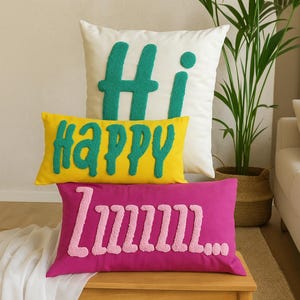 Puede incluir: Tres cojines decorativos con texto bordado. El cojín superior es blanco con "Hi" en turquesa. El cojín central es amarillo con "happy" en turquesa. El cojín inferior es magenta con "lmmmmm..." en rosa.