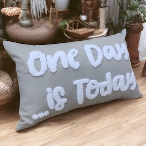 Può includere: Un cuscino grigio con lettere bianche e soffici che recitano "One Day is Today".