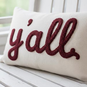 May include: Cream-coloured rectangular cushion with the word "y'all" embroidered in a burgundy script. The cushion has a textured surface and is placed on a white wooden bench. The embroidered letters are raised, adding dimension to the cushion.