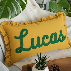 Può includere: Cuscino rettangolare giallo senape con testo ricamato verde "Lucas" e bordo a balze. Il cuscino è su una superficie colorata chiara con piante verdi e frondose sullo sfondo. Una piccola pianta succulenta in un vaso bianco è in primo piano.