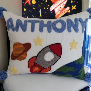 Op de afbeelding: Een wit kussen met blauwe geborduurde letters die "ANTHONY" zeggen. Het kussen heeft een raket, een planeet en sterren geborduurd.