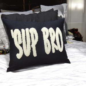 Peut inclure: Un coussin décoratif noir avec les mots "SUP BRO" en grandes lettres blanches texturées. Le coussin rectangulaire est posé sur un lit blanc, avec d'autres coussins en arrière-plan.