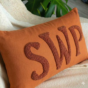 May include: A rectangular rust-coloured throw cushion featuring the letters SWP in a raised, textured, punch needle embroidery style. The letters are in a matching rust tone, creating a monochromatic look on the smooth fabric surface of the cushion.