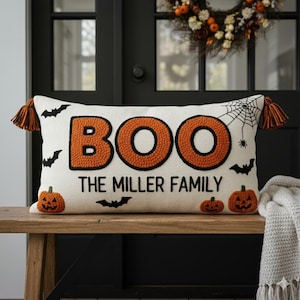 May include: Cream-coloured rectangular Halloween cushion with the word "BOO" in orange letters with black outlines. The text "THE MILLER FAMILY" is below. It features black bats, pumpkins, a spider web, and orange tassels.