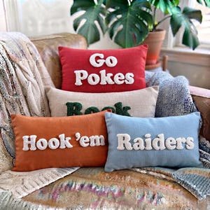 May include: A collection of four rectangular throw pillows with textured white lettering. The top pillow is red with 'Go Pokes', the second is beige with green text, the third is orange with 'Hook 'em', and the bottom is light blue with 'Raiders'.