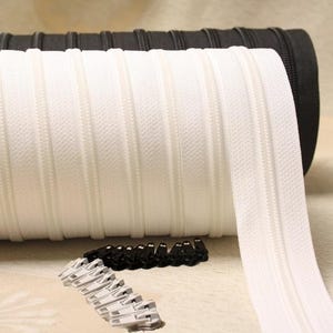 May include: A roll of white zipper tape with black zipper pulls. The zipper tape is coiled and the zipper pulls are scattered on a white surface.
