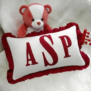 May include: A rectangular white throw pillow featuring a dark red ruffled border and the letters ASP embroidered in a textured dark red font. A plush teddy bear with white and red fur is positioned directly behind the pillow.