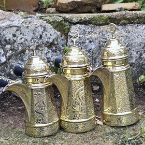 May include: Three ornate, gold-colored coffee pots with intricate embossed designs. Each pot has a curved spout, a handle, and a decorative lid with a star-shaped finial. The pots are arranged on a mossy stone surface, with greenery in the background.