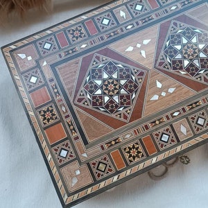 Puede incluir: Una caja de madera con intrincados patrones geométricos incrustados en tonos marrones, rojos y blancos. La caja tiene una tapa con bisagras y está decorada con detalles de nácar.