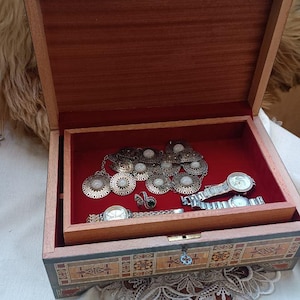 Puede incluir: Caja de joyería de madera abierta con interior de terciopelo rojo. La caja contiene collares, anillos y relojes. El exterior está decorado con patrones incrustados. La caja está abierta, revelando el contenido.