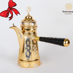 May include: A gold-colored metal coffee pot with a black handle and intricate floral designs. The pot has a crescent moon and star design on the lid. The text "Oriental Touches" is visible in the upper right corner of the image.