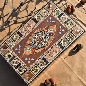 Puede incluir: Una caja rectangular de madera con intrincados patrones geométricos incrustados en tonos marrón, beige, rojo y blanco. La caja presenta un diseño ovalado central y bordes decorativos. Bellotas y ramitas están esparcidas alrededor.