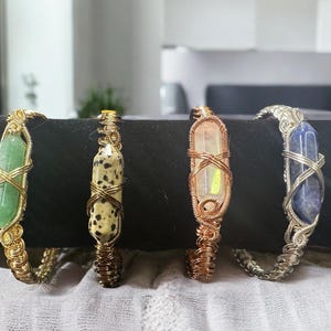 May include: Four wire-wrapped crystal bracelets. The crystals are green, speckled, iridescent, and blue. The wire is gold, copper, and silver. The bracelets are displayed on a black surface.