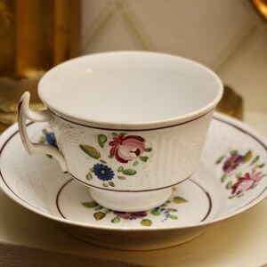 May include: A white teacup and saucer with a floral pattern in shades of pink, red, blue, and green. The cup has a handle and a textured surface. The saucer has a matching floral pattern.